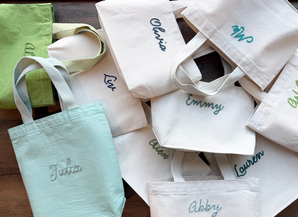 A collection of personalized canvas tote bags in various colors, including cream and light green, featuring embroidered names like Julia, Emmy, and Lauren.