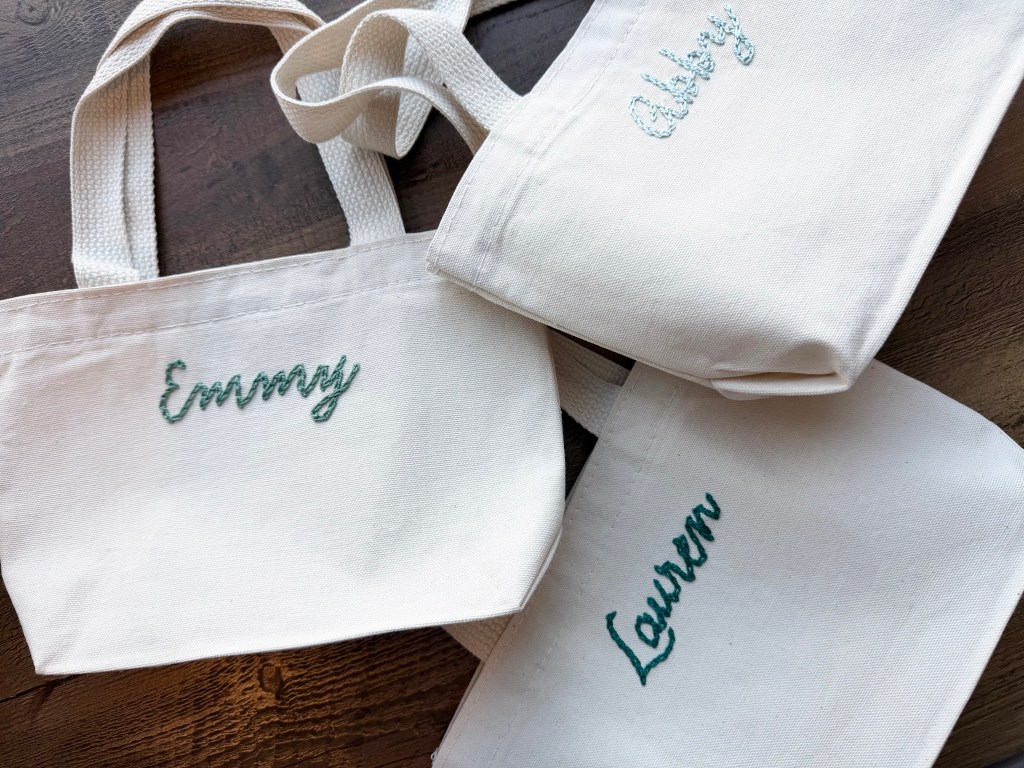Three personalized cream-colored tote bags with embroidered names 'Emmy', 'Abby', and 'Lauren' in green and blue threads.