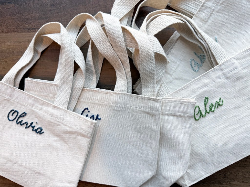 A collection of white canvas tote bags featuring embroidered names 'Olivia', 'Cass', and 'Alex' in different colors.